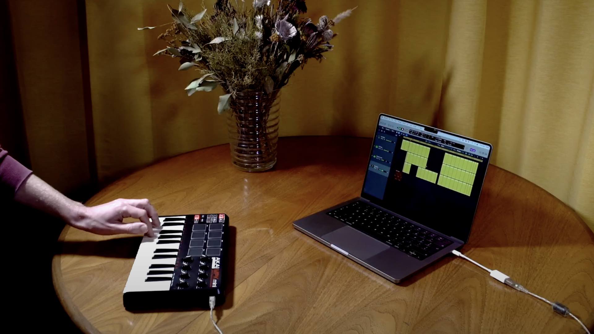 A hand playing a small keyboard attached to a laptop on a wooden table with a bunch of flowers on it